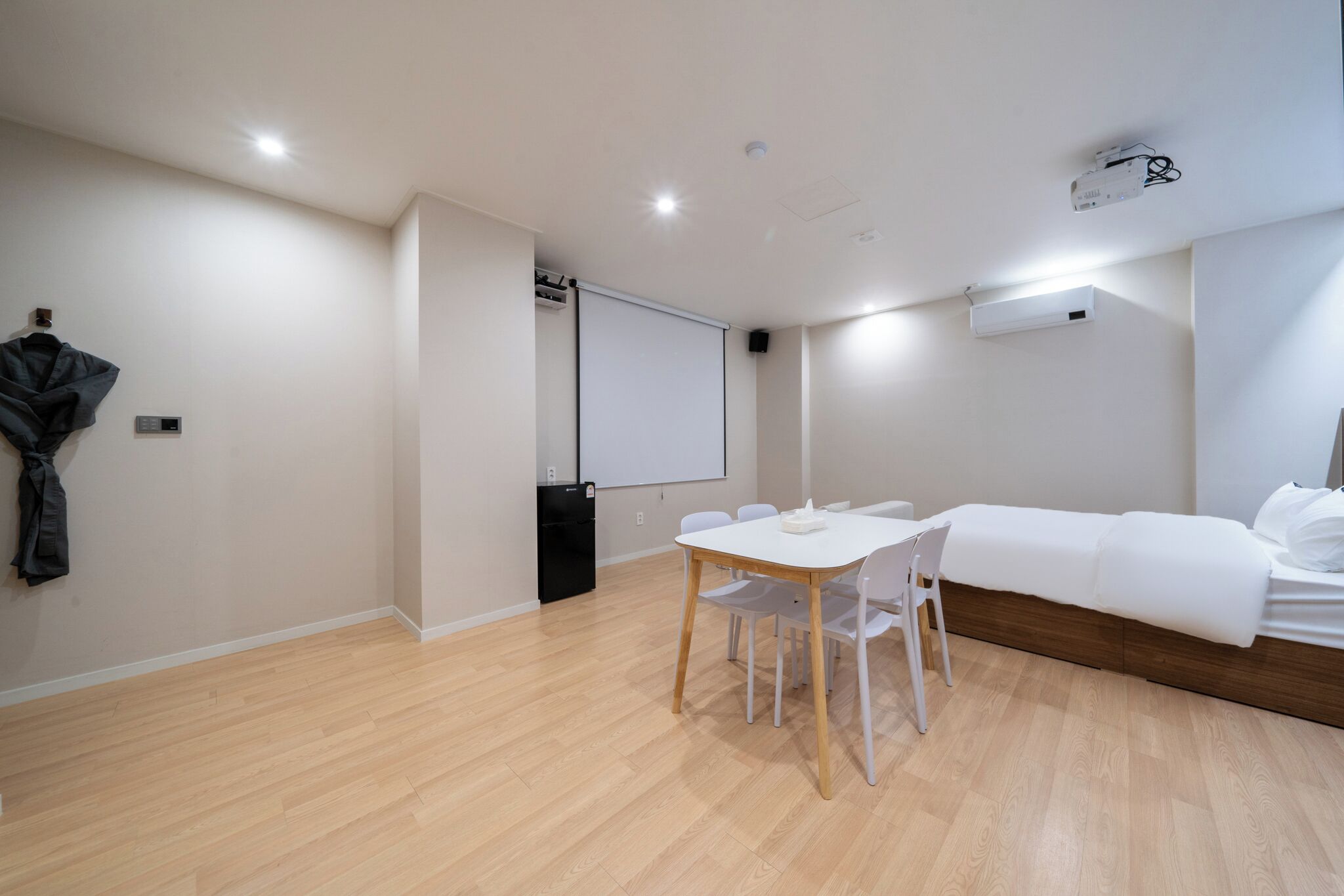 Une chambre d’hôtel moderne avec un plancher en bois, un lit blanc, une petite table avec des chaises, un écran de projection et un porte-manteau.