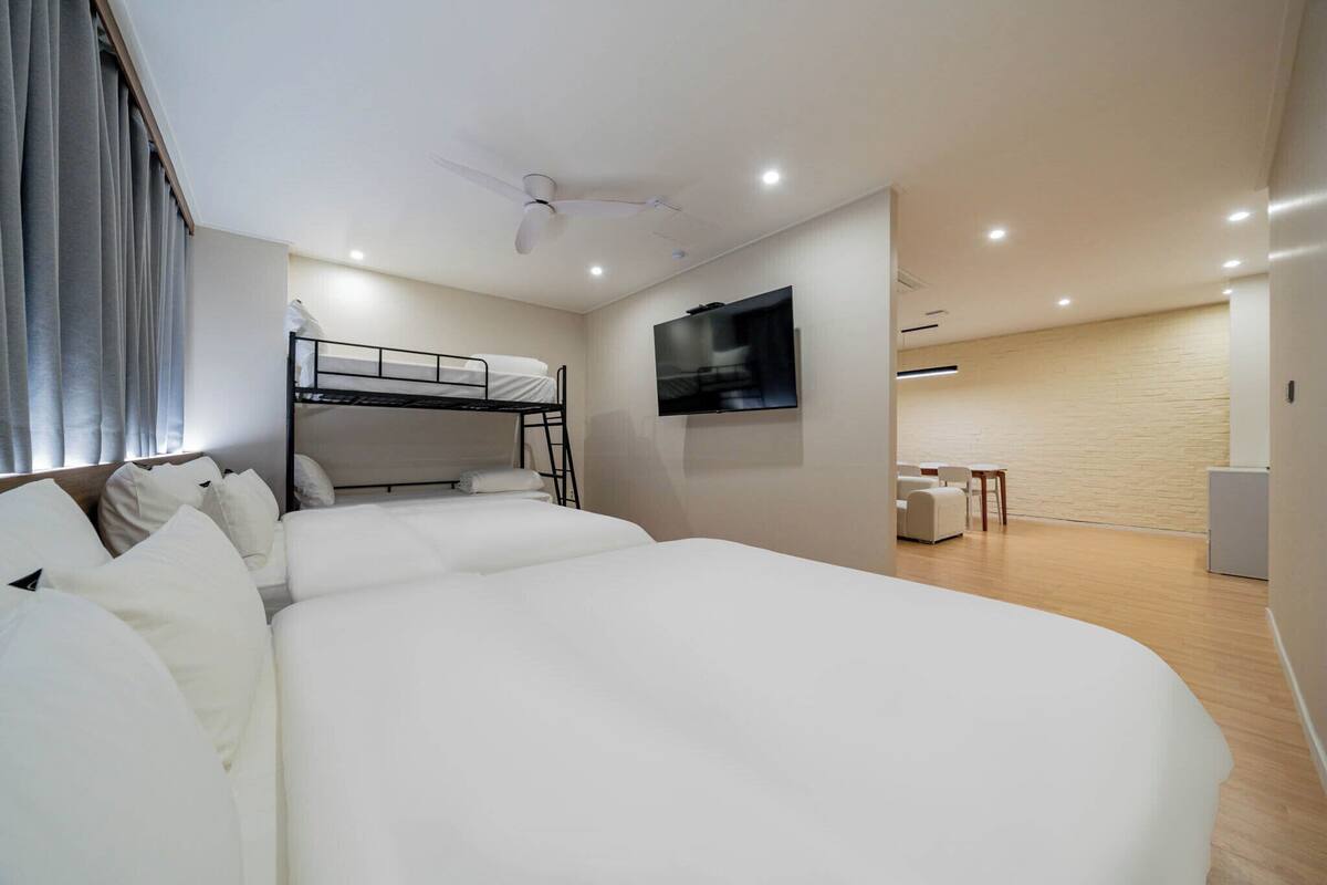 Une chambre d’hôtel moderne équipée d’un lit superposé, d’un téléviseur à écran plat fixé au mur, d’un ventilateur de plafond et d’un petit coin salon avec une table et des chaises.