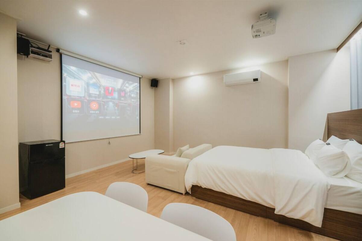 Une chambre d’hôtel moderne équipée d’un lit, d’un petit canapé, d’un bureau et d’un écran de projection diffusant une vidéo.