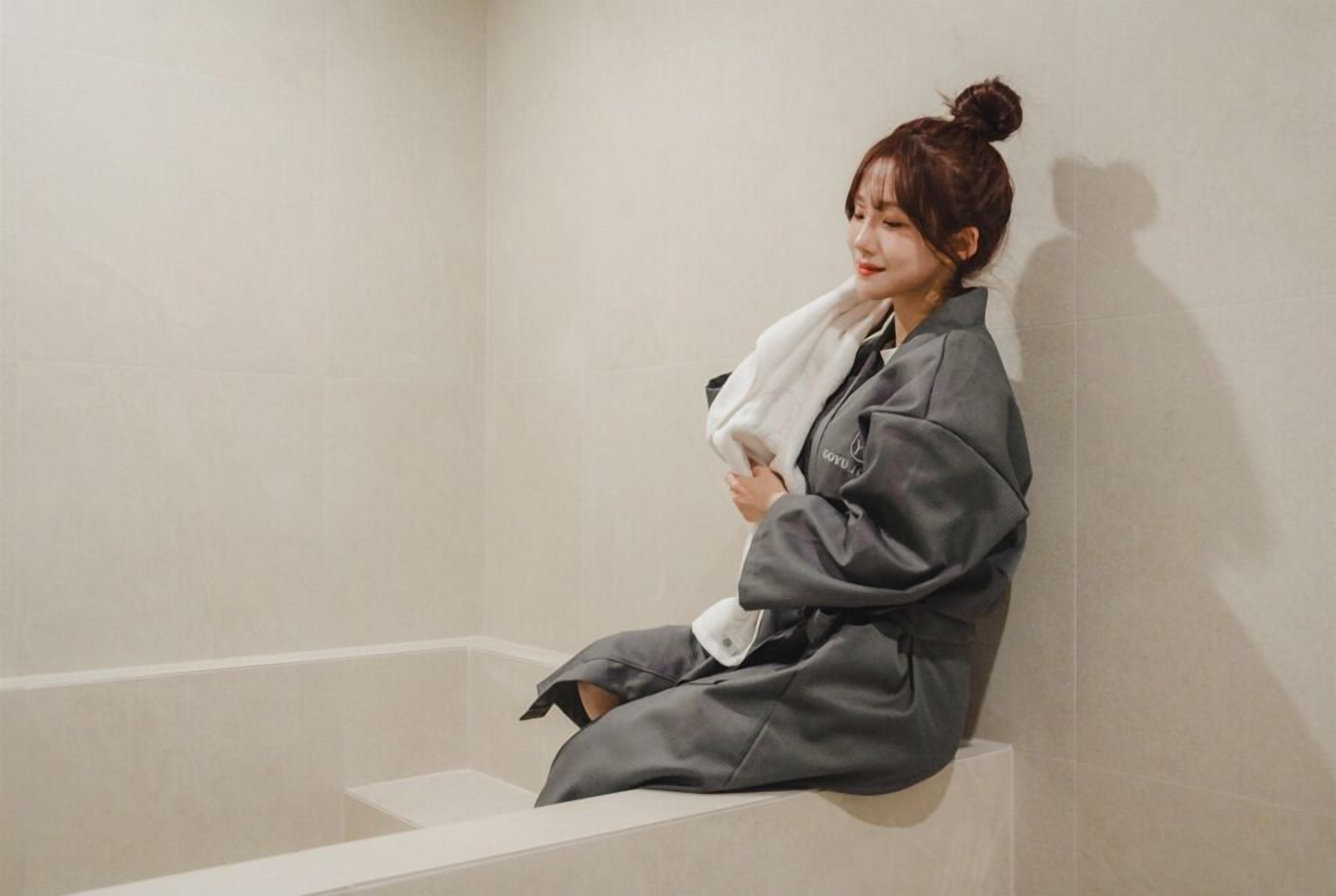 A person sitting on a bathtub step in a bathroom with a plain wall and a white floor.