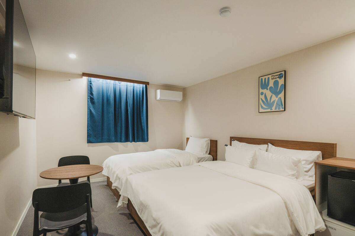 Une chambre d’hôtel avec deux lits, une petite table, une chaise, un téléviseur et une fenêtre avec des rideaux.