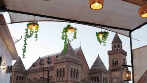 A covered outdoor area with wicker hanging lamps, a red and white patterned wall, and a view of a historic building with a clock tower.