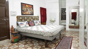 A bedroom with a patterned bedspread, a wooden headboard, a red curtain, a small table with a vase, and a tiled floor.