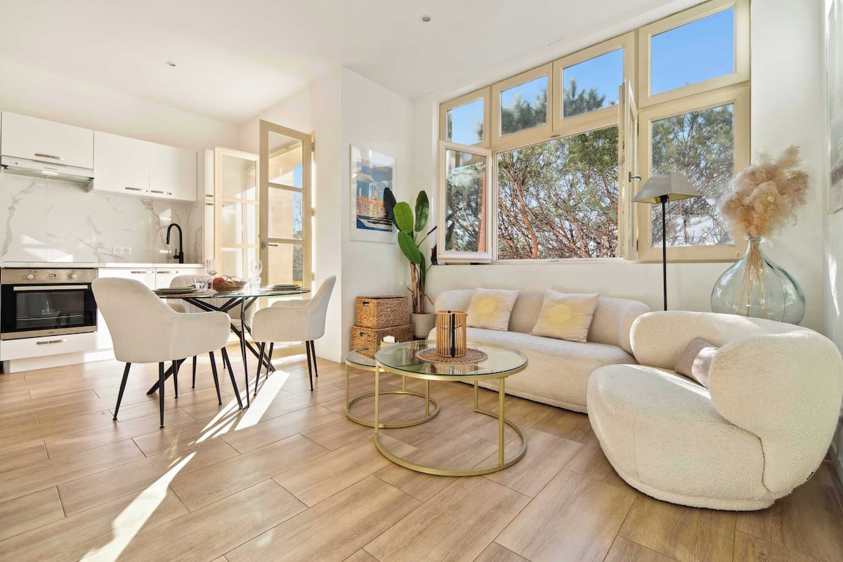 Un salon moderne avec un canapé, des fauteuils et une table basse. Il y a également un coin cuisine avec des placards et une cuisinière. De grandes fenêtres laissent entrer la lumière naturelle et offrent une vue sur les arbres.