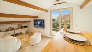 Una sala de estar moderna con techo de madera, un televisor de pantalla plana, una mesa de comedor con platos y cubiertos, y vista a edificios a través de un ventanal grande.