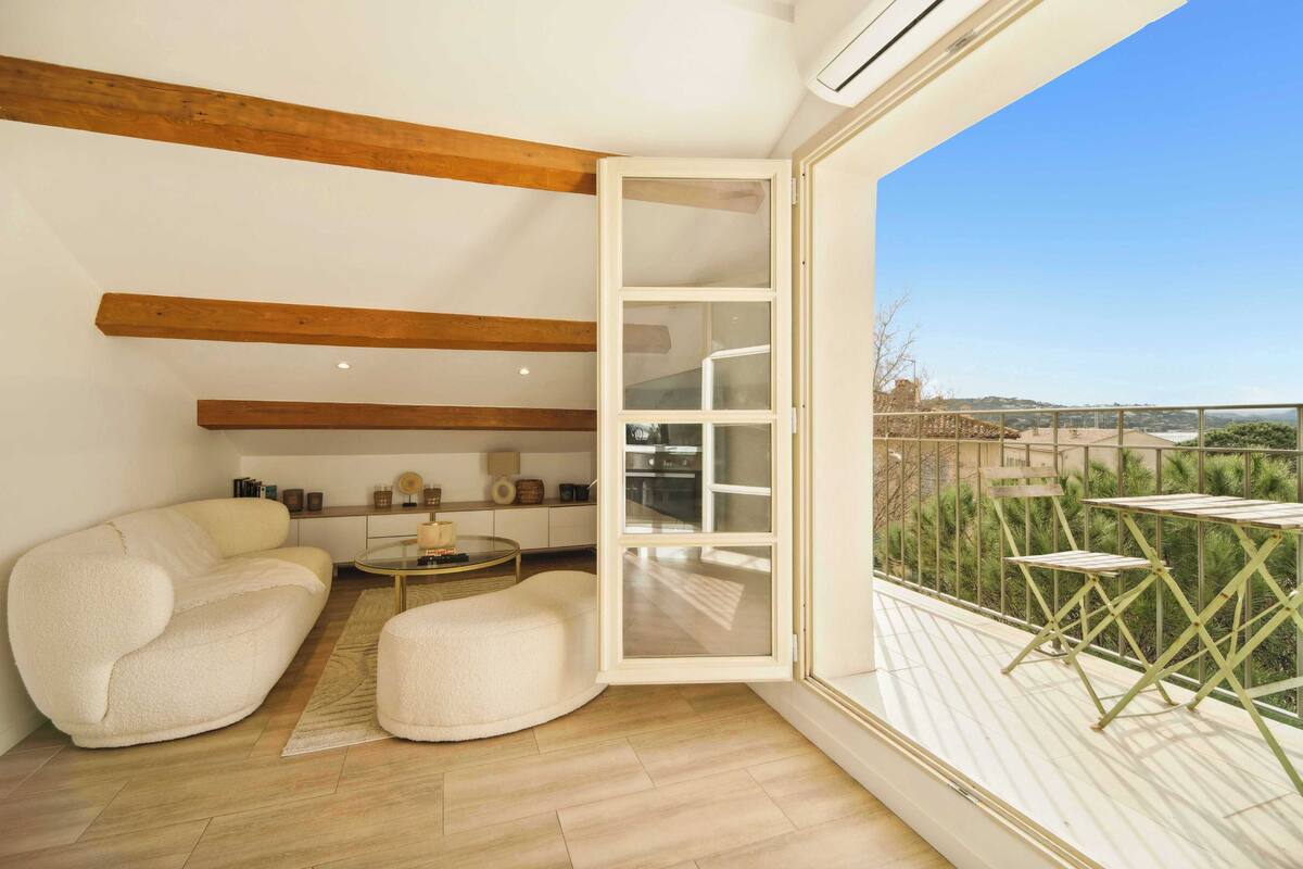 Un salon moderne avec un canapé, un pouf et une porte vitrée donnant sur un balcon aménagé avec du mobilier d’extérieur.