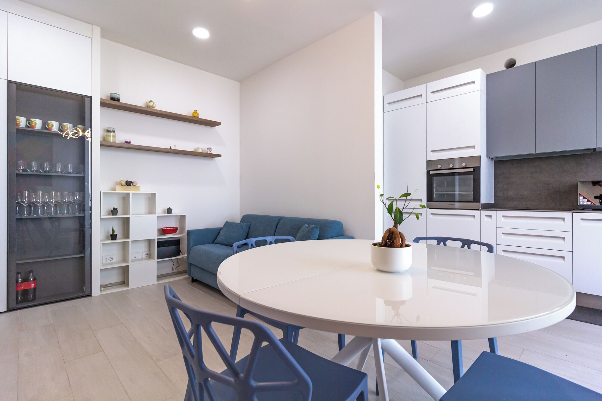A modern kitchen with white cabinets, a built-in oven, and a dining area with a round table and blue chairs.