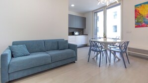 A modern living room with a blue sofa, a dining table with chairs, and a kitchen in the background.