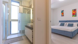 A modern bedroom with a bed, a bedside table, and a nightstand. There is a bathroom with a sink, mirror, and glass shower enclosure. The room has light-colored walls and a wooden floor.