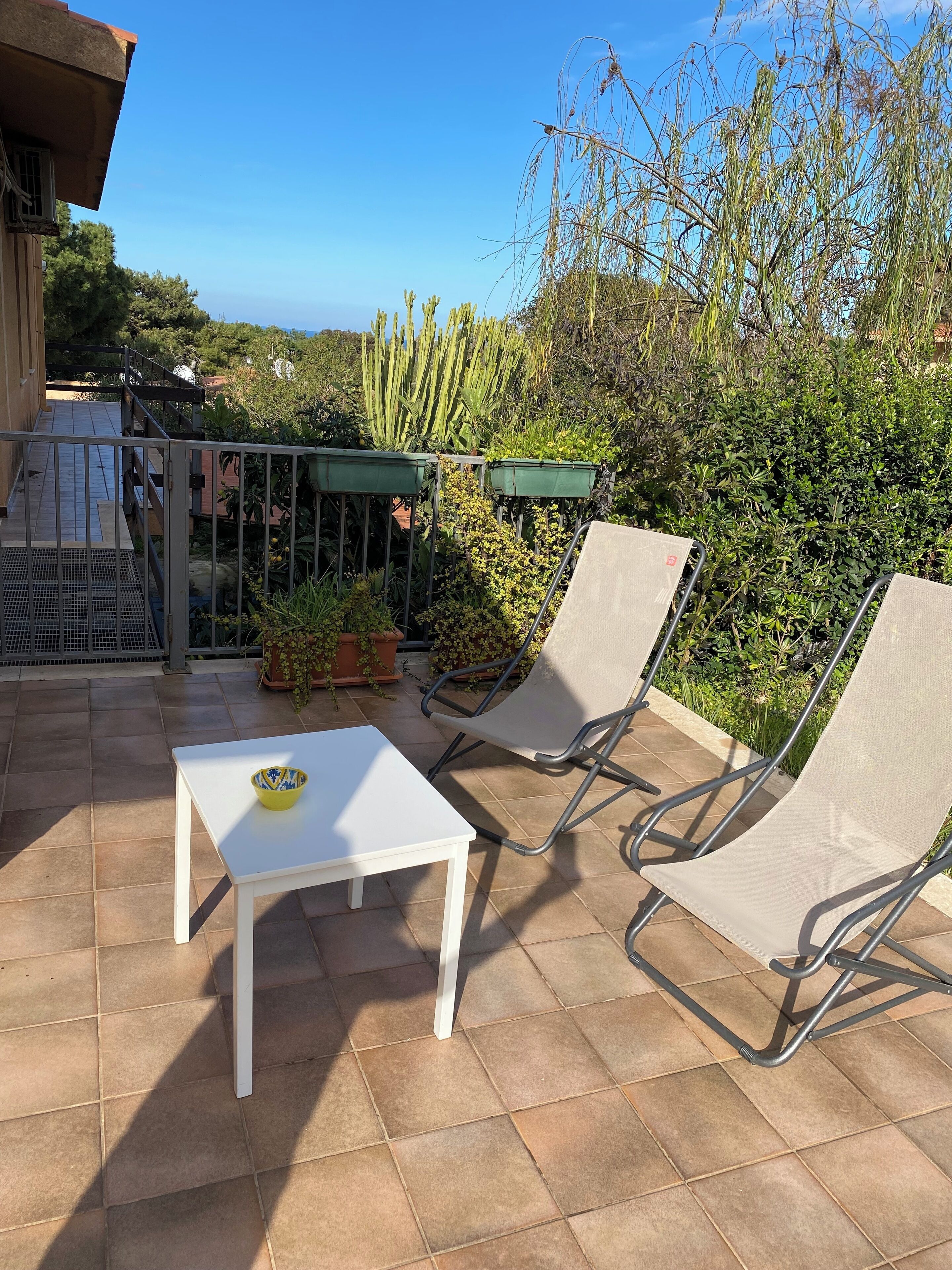 An outdoor patio with a white table and two lounge chairs, surrounded by greenery and a metal fence.