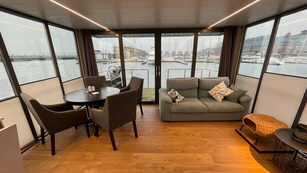 Une pièce moderne avec un plancher en bois, une table ronde entourée de chaises, un canapé gris agrémenté de coussins à motifs et de grandes fenêtres offrant une vue sur les bateaux et les bâtiments.