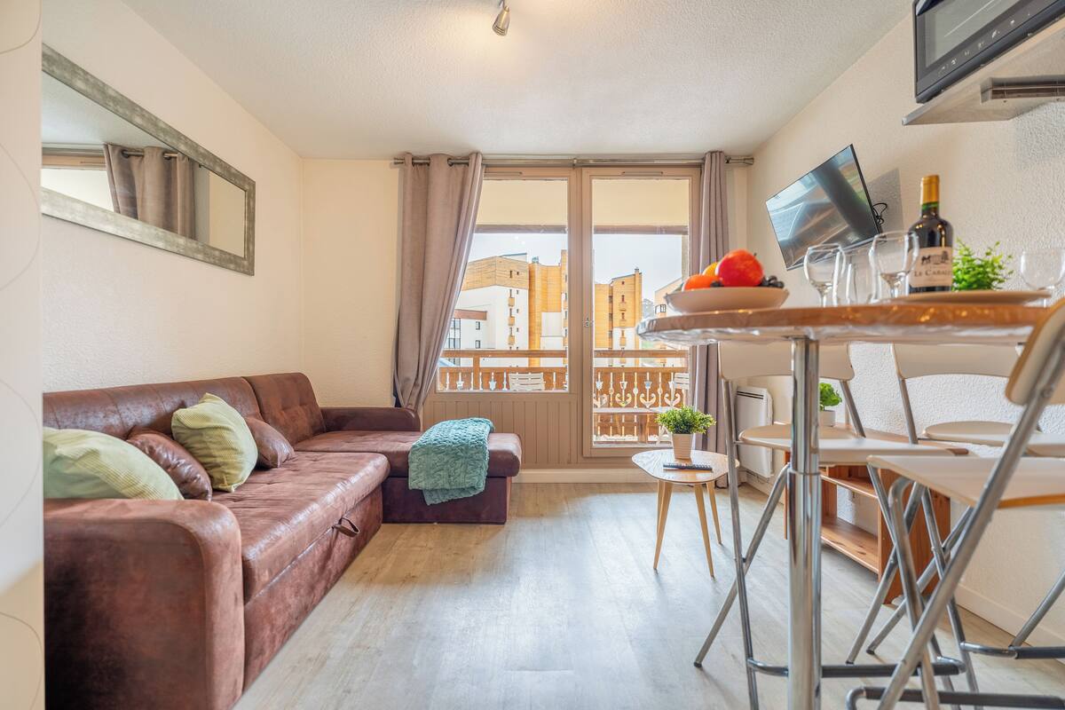 A modern living room with a brown leather sofa, a small round table with a bottle and glasses, and a TV on the wall.
