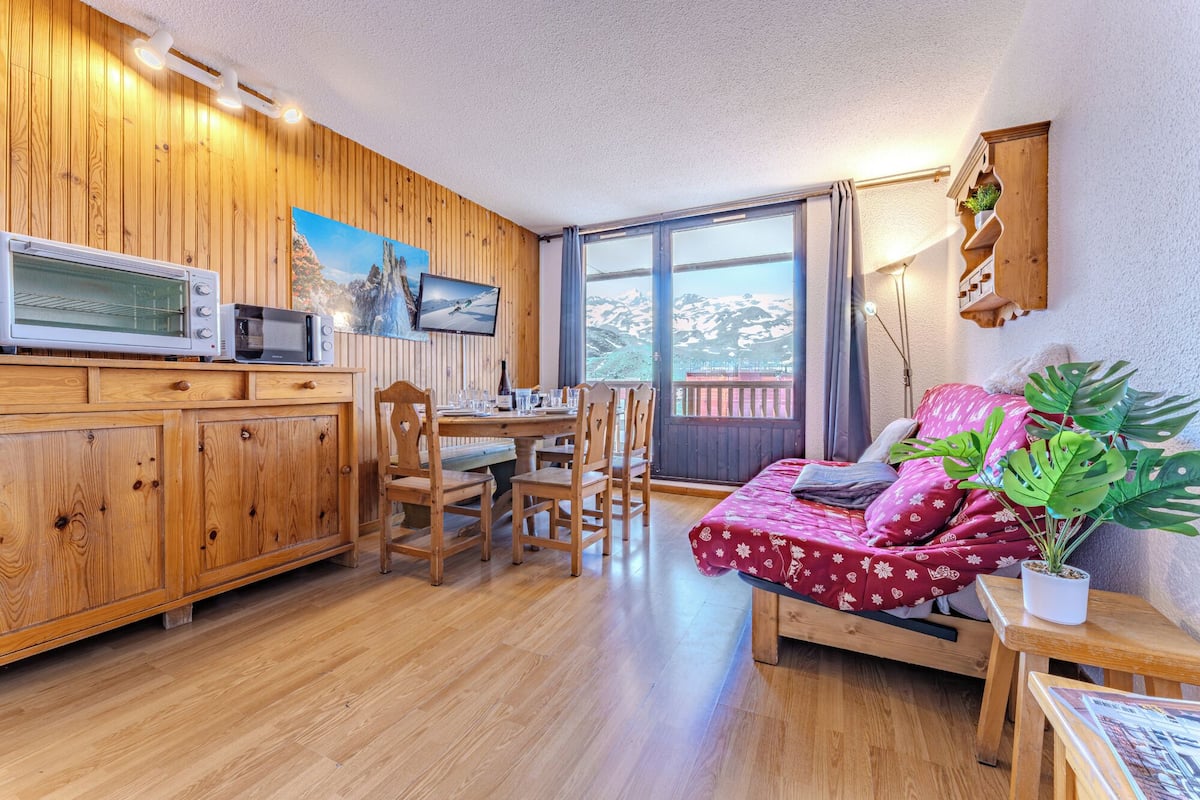 A cozy room with wooden walls, a dining area with chairs, a bed with a red floral bedspread, and a large window with a mountain view.