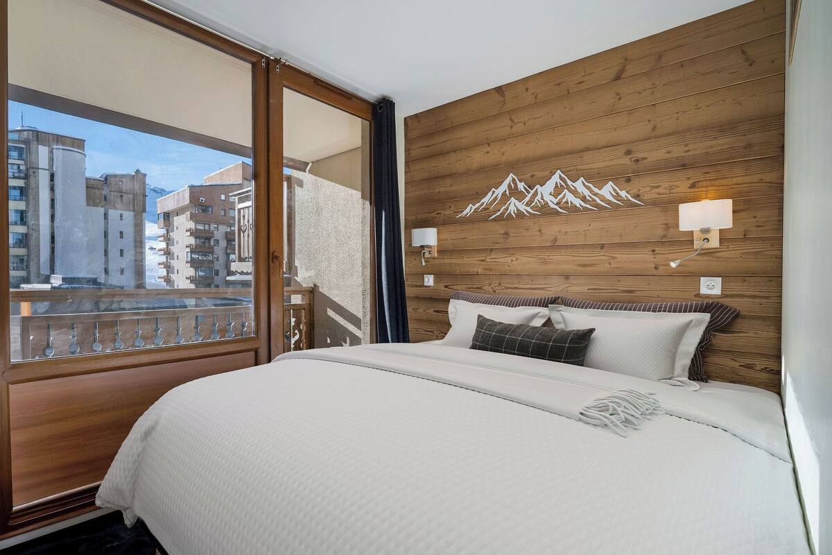 A hotel room with a large bed, wooden panel walls, a mountain mural, and a balcony with a city view.