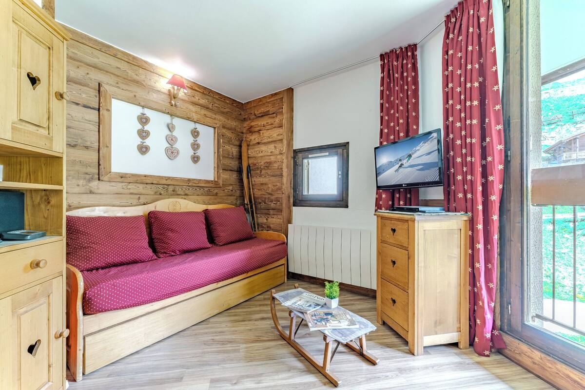 A cozy room with a wooden bed, a small table with a plant, a TV, and a window with curtains.