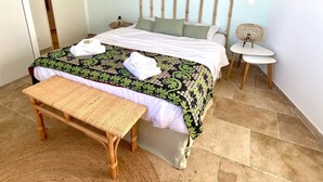 Een netjes opgemaakt bed met een bedsprei met patroon, een nachtkastje met een lamp, een klein rond tafeltje met een stoel en een bankje voor het bed.