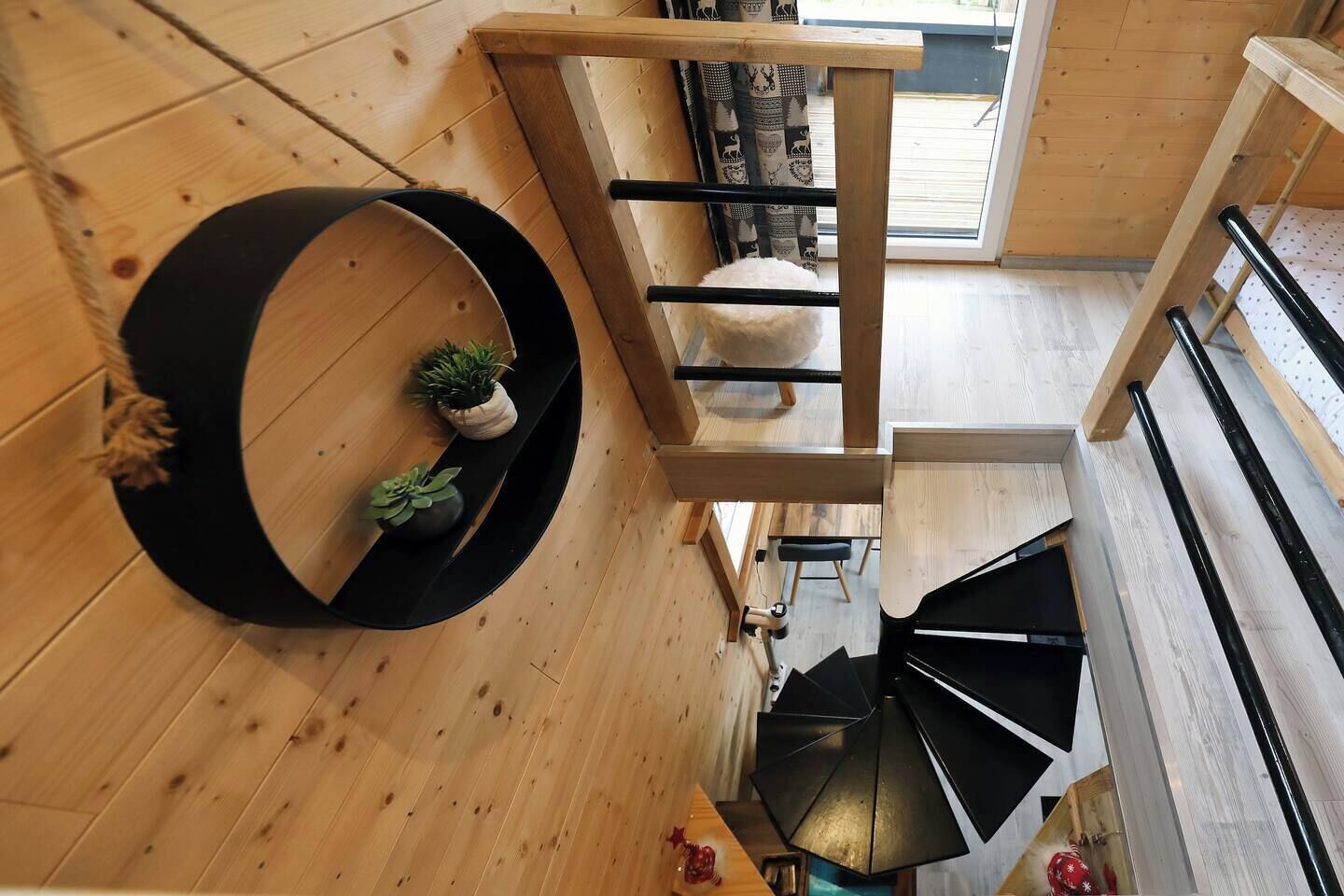 Un intérieur en bois avec un escalier en colimaçon, une étagère circulaire noire ornée de plantes et une vue sur l’extérieur par une fenêtre.