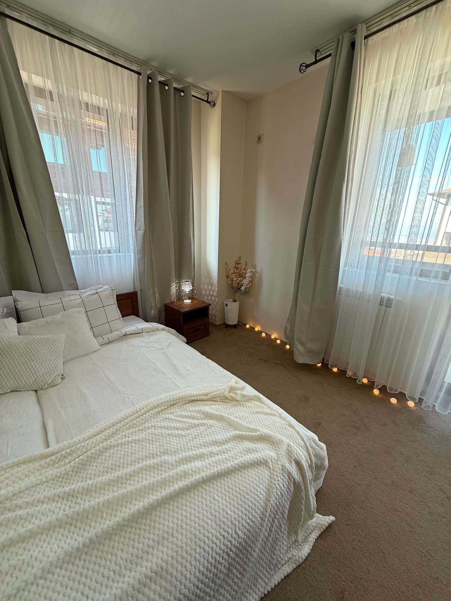 Een slaapkamer met een groot bed, lichtgordijnen, een nachtkastje en een vaas met bloemen.