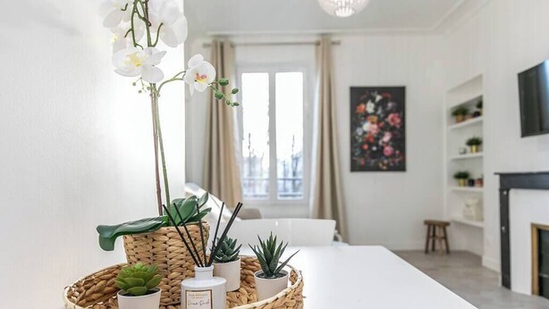 Een moderne woonkamer met een witte tafel, een geweven dienblad met planten en kaarsen, een groot raam met gordijnen, en een schilderij aan de muur.