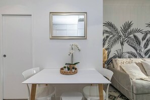 Une chambre moderne avec une table blanche, des chaises, un miroir et un canapé agrémenté d’un papier peint aux motifs tropicaux.
