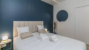Een keurig opgemaakt bed met witte lakens en opgerolde handdoeken, een nachtkastje met een lamp, en een ronde spiegel aan de muur.