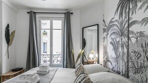 Una habitación de hotel con cama, ventana con cortinas, un espejo y un mural de plantas tropicales en la pared.