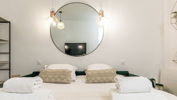 A hotel room with a large round mirror, two bedside tables, a flat-screen TV, and a bed with white linens and patterned pillows. - Splendide Appart 2P- Popincourt Paris centre (Paris)