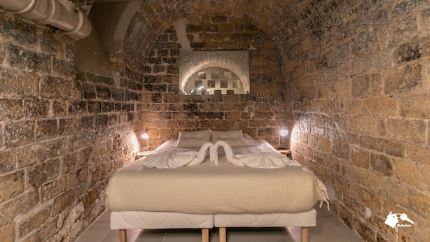 Una habitación con paredes de piedra, una cama, dos lámparas de noche y una mesita.