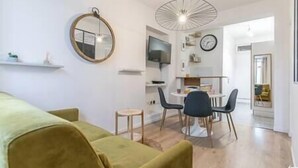 Un salon moderne avec un canapé vert, un miroir rond, un coin repas comprenant une petite table et des chaises, et un téléviseur fixé au mur.