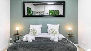 A neatly made bed with a dark patterned bedspread, two white pillows with a green design, and a large mirror above the bed. There are two bedside tables with lamps on either side of the bed.
