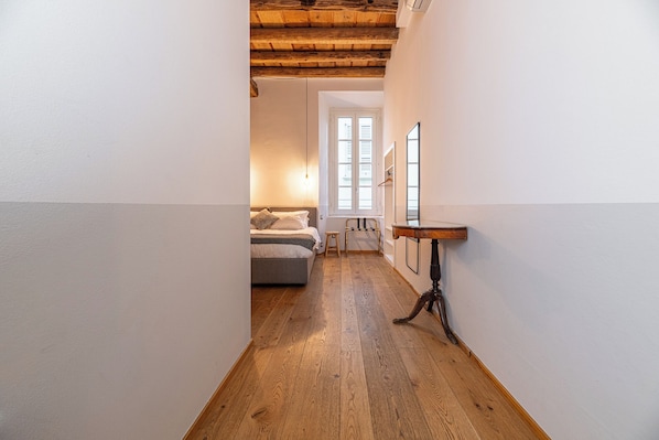 A narrow hallway with wooden flooring, a bed with a headboard, a small table, and a window with shutters.