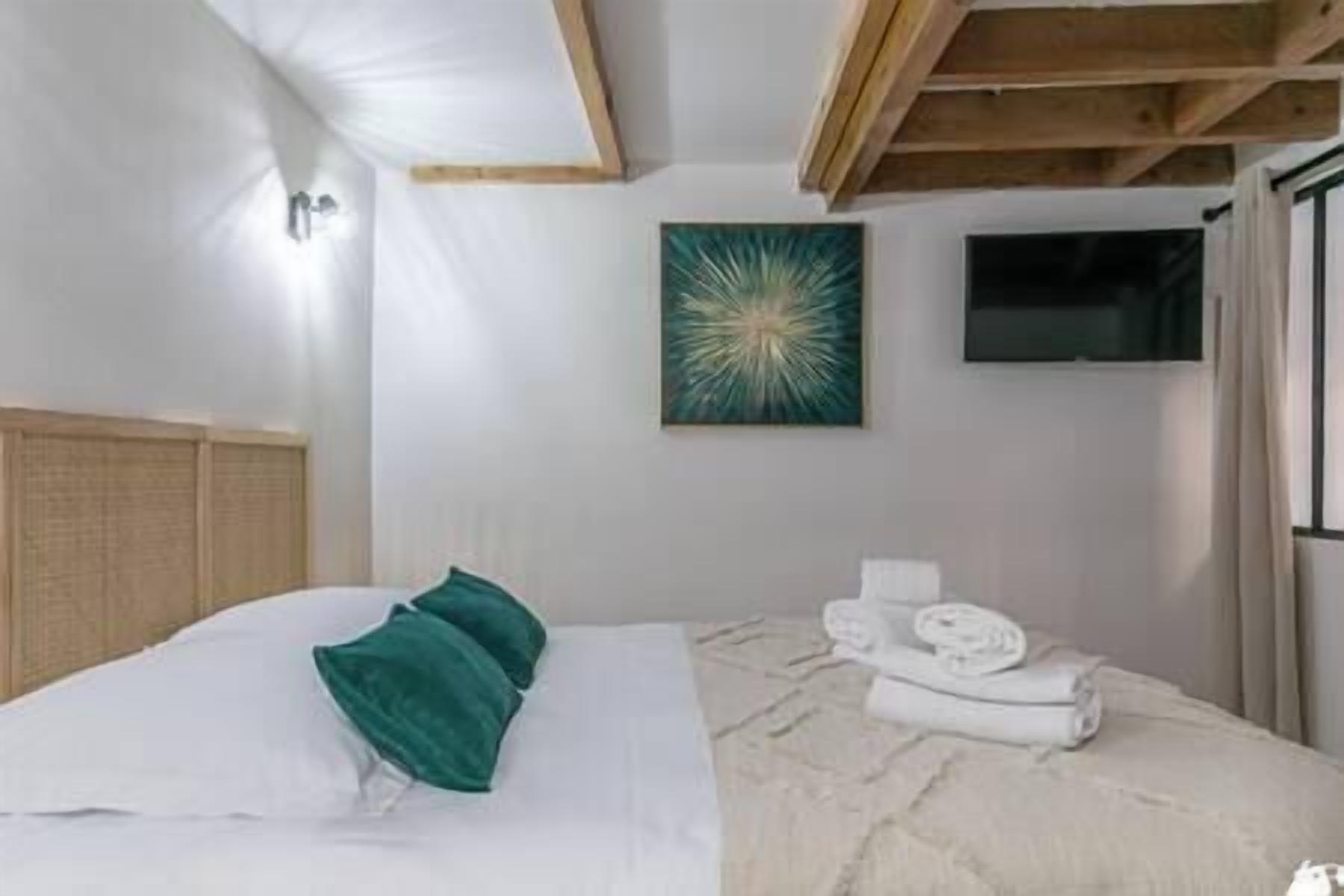 A bedroom with a bed, a green pillow, a white blanket, a towel, and a painting on the wall.