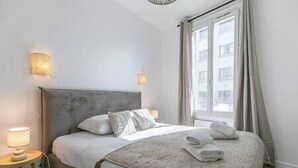A neatly made bed with a grey headboard, a bedside lamp, and a window with curtains. - MBA - Fontenay - Splendide Appart Proche Vincennes (Fontenay-sous-Bois)