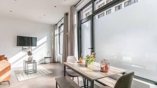 A modern, well-lit room with a large window, a wooden table with chairs, a flat-screen TV, and a small side table with a vase of flowers. - MBA - Fontenay - Splendide Appart Proche Vincennes (Fontenay-sous-Bois)