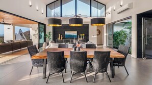 A spacious dining area with a large wooden table and black woven chairs, a modern kitchen with a view, and a high ceiling with hanging lights.