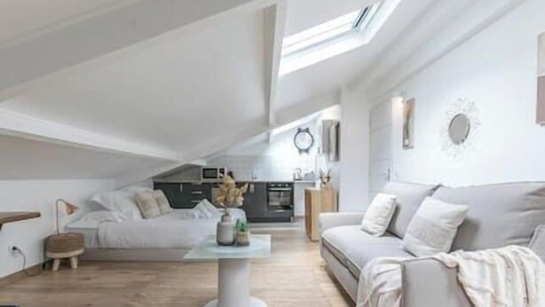 Ein großzügiger Loft mit hoher Decke, einem Oberlicht, einer gemütlichen Sitzecke mit Sofa und Sessel, einem kleinen runden Tisch und einer Küche im Hintergrund.