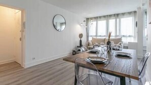 A modern dining area with a wooden table set for four, a round mirror on the wall, and a large window providing natural light. - MBA - Splendide Appart Proche Château Vincennes (Vincennes)