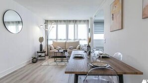 A modern living room with a sofa, a dining table set for four, and a large mirror on the wall. - MBA - Splendide Appart Proche Château Vincennes (Vincennes)