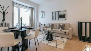 A modern living room with a sofa, a dining table, and chairs. There is a large window providing natural light.