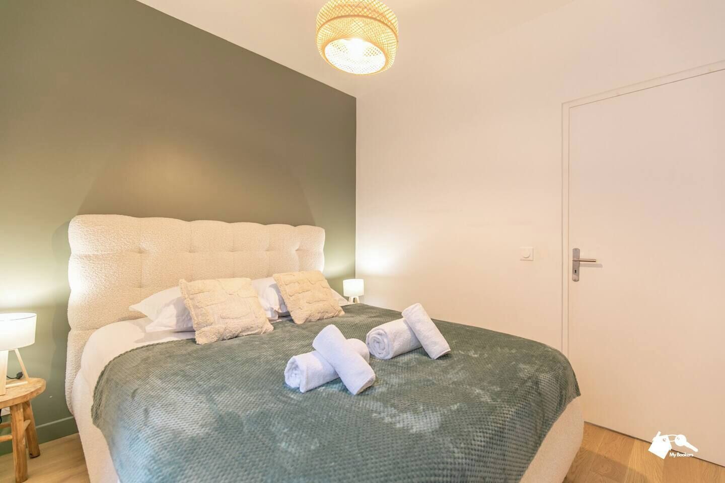 A neatly made bed with a green bedspread and white pillows, a bedside lamp, and a door to the right.
