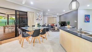 Eine moderne Küche mit einer zentralen Kochinsel, einem Essbereich mit Glastisch sowie einem Wohnbereich mit Sofa und Fernseher.