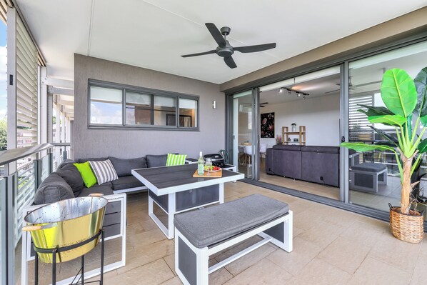 A modern outdoor living area with a sofa, dining table, and bench, a ceiling fan, and a large sliding glass door leading to an adjacent room. - Waterfront Monsoon Suite: Top Floor 2 Bedroom Apt (Darwin City)