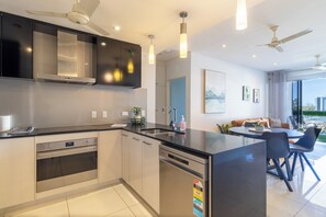 Une cuisine moderne dotée d’un îlot central, d’un four encastré et d’un lave-vaisselle. On y trouve également un coin repas avec une table et des chaises, ainsi qu’un ventilateur de plafond.