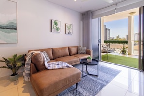 Un salon moderne avec un canapé en cuir marron, une petite table ronde et une plante en pot. Deux tableaux encadrés ornent le mur, et une grande porte-fenêtre coulissante donne accès à un espace extérieur avec vue sur les bâtiments.