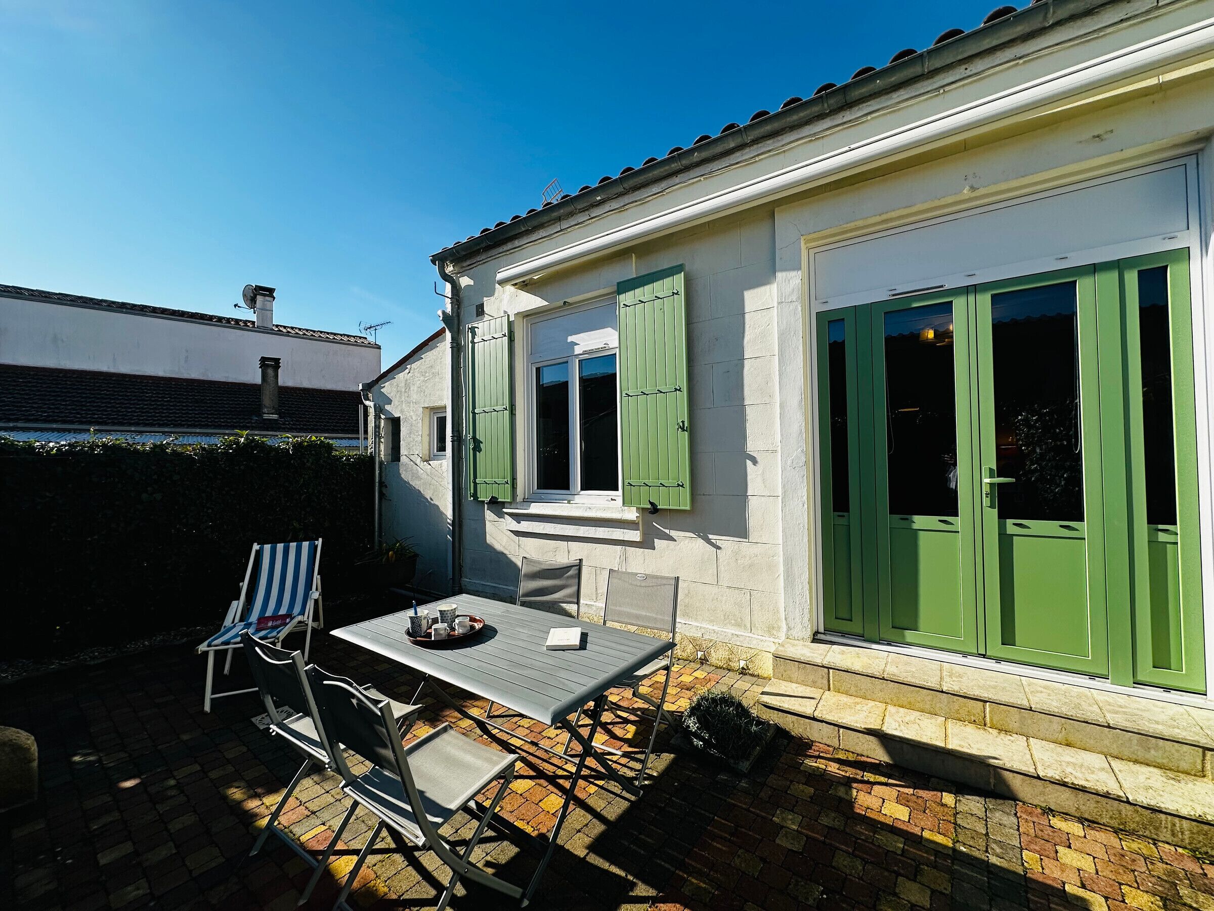 Een terras met een tafel en stoelen, een kleine tuin en een gebouw met groene luiken.
