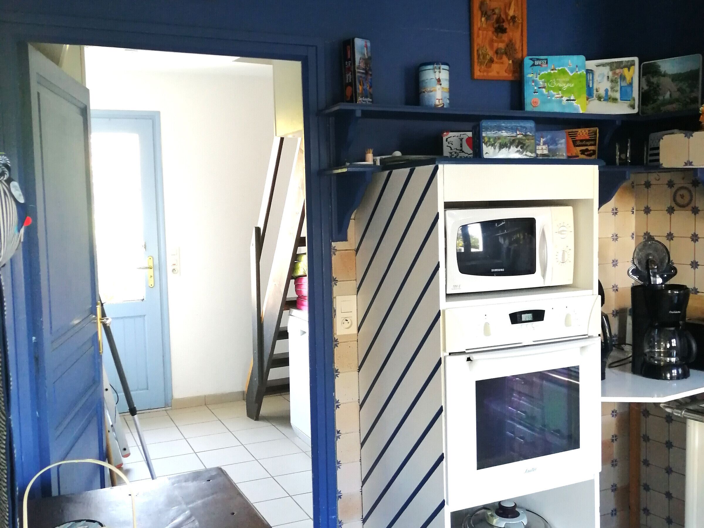 Een kleine keuken met een magnetron, oven en koffiezetapparaat. Een ladder staat tegen de muur, en een deur leidt naar een andere kamer.