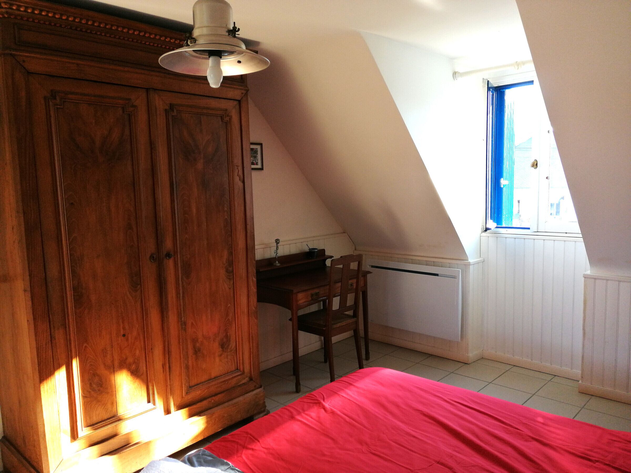 Une chambre à coucher avec une armoire en bois, un bureau, une chaise et un lit recouvert d’une couverture rouge.