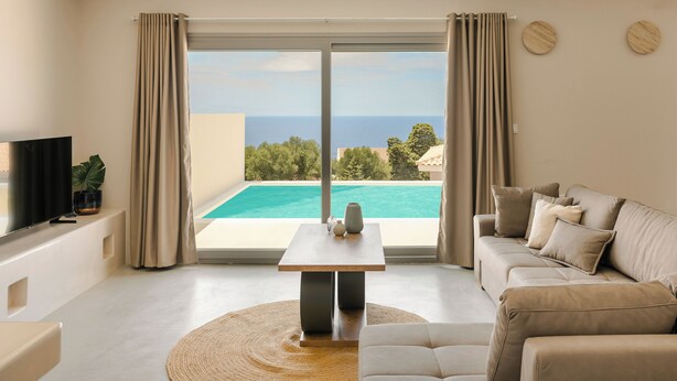 A modern living room with a large window, a pool view, a flat-screen TV, and a beige sofa. - Villa Armonia - Zen Villas (Fiskardo)