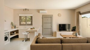 A modern living room with a sofa, a TV, and a kitchen area with a window and an air conditioning unit. - Villa Armonia - Zen Villas (Fiskardo)