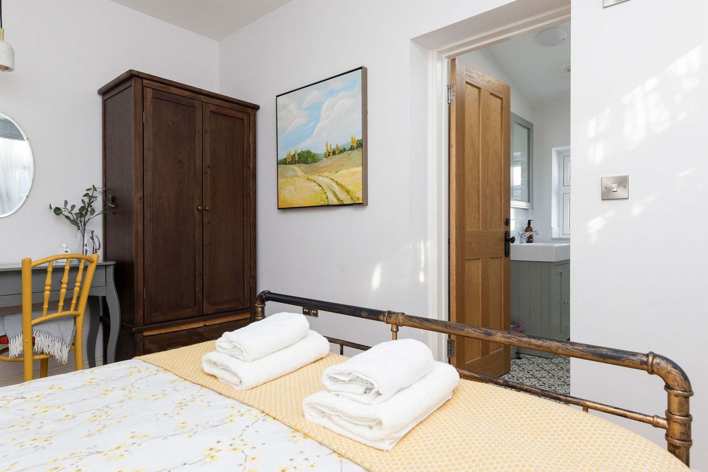 Una camera da letto con un letto, un armadio in legno, un quadro, uno specchio e un bagno visibile attraverso una porta aperta.